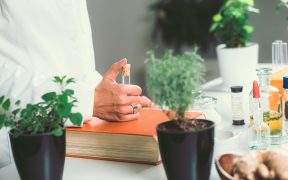 kobieta w biąłym kitlu trzyma w dłoni szklaną fiolkę, na stole stoją rośliny i inne preparaty w szklanych menzurkach